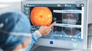 English: "An eye care professional in a white coat pointing at a retinal scan displayed on a computer monitor, showing an orange-colored fundus image of the back of an eye with targeting crosshairs, while a patient wearing a face mask is visible in the background.