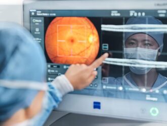 English: "An eye care professional in a white coat pointing at a retinal scan displayed on a computer monitor, showing an orange-colored fundus image of the back of an eye with targeting crosshairs, while a patient wearing a face mask is visible in the background.