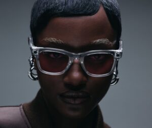 Portrait en gros plan d'un homme noir portant des lunettes de soleil distinctives à monture transparente avec des verres foncés et des éléments métalliques sur les côtés. Il a les cheveux courts et foncés ainsi qu'une barbe, photographié sur un fond gris neutre avec un éclairage dramatique.