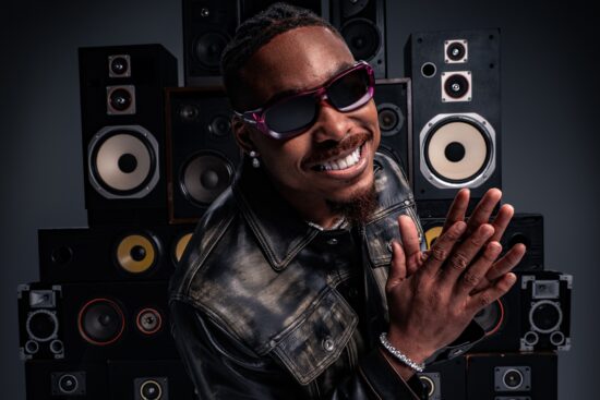 Justin Jefferson en veste de cuir noir et lunettes de soleil dégradé violet souriant et saluant devant un mur de haut-parleurs vintage
