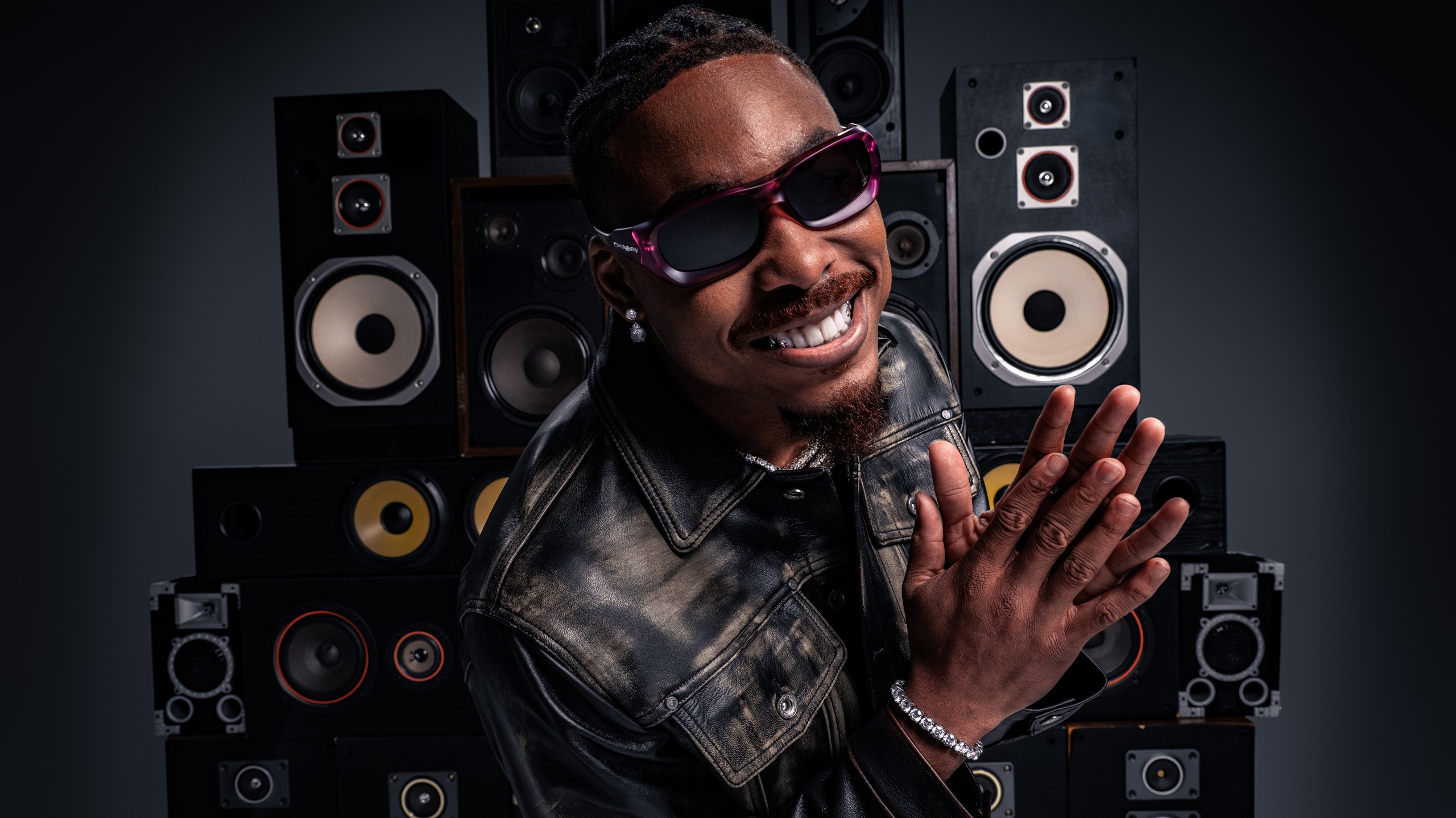 Justin Jefferson en veste de cuir noir et lunettes de soleil dégradé violet souriant et saluant devant un mur de haut-parleurs vintage