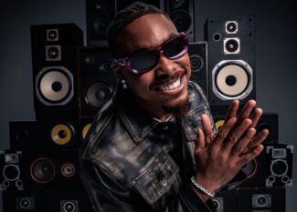 Justin Jefferson in black leather jacket and purple gradient sunglasses smiling and waving at camera in front of wall of vintage speakers