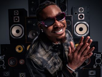 Justin Jefferson in black leather jacket and purple gradient sunglasses smiling and waving at camera in front of wall of vintage speakers
