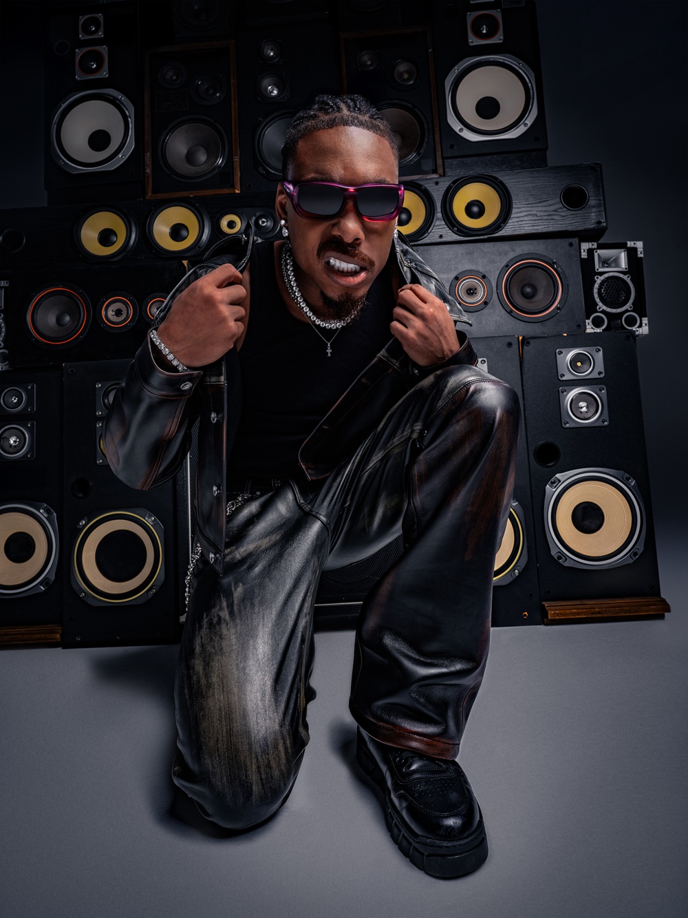 Justin Jefferson in black leather jacket and purple gradient sunglasses adjusting his collar while kneeling in front of wall of vintage speakers