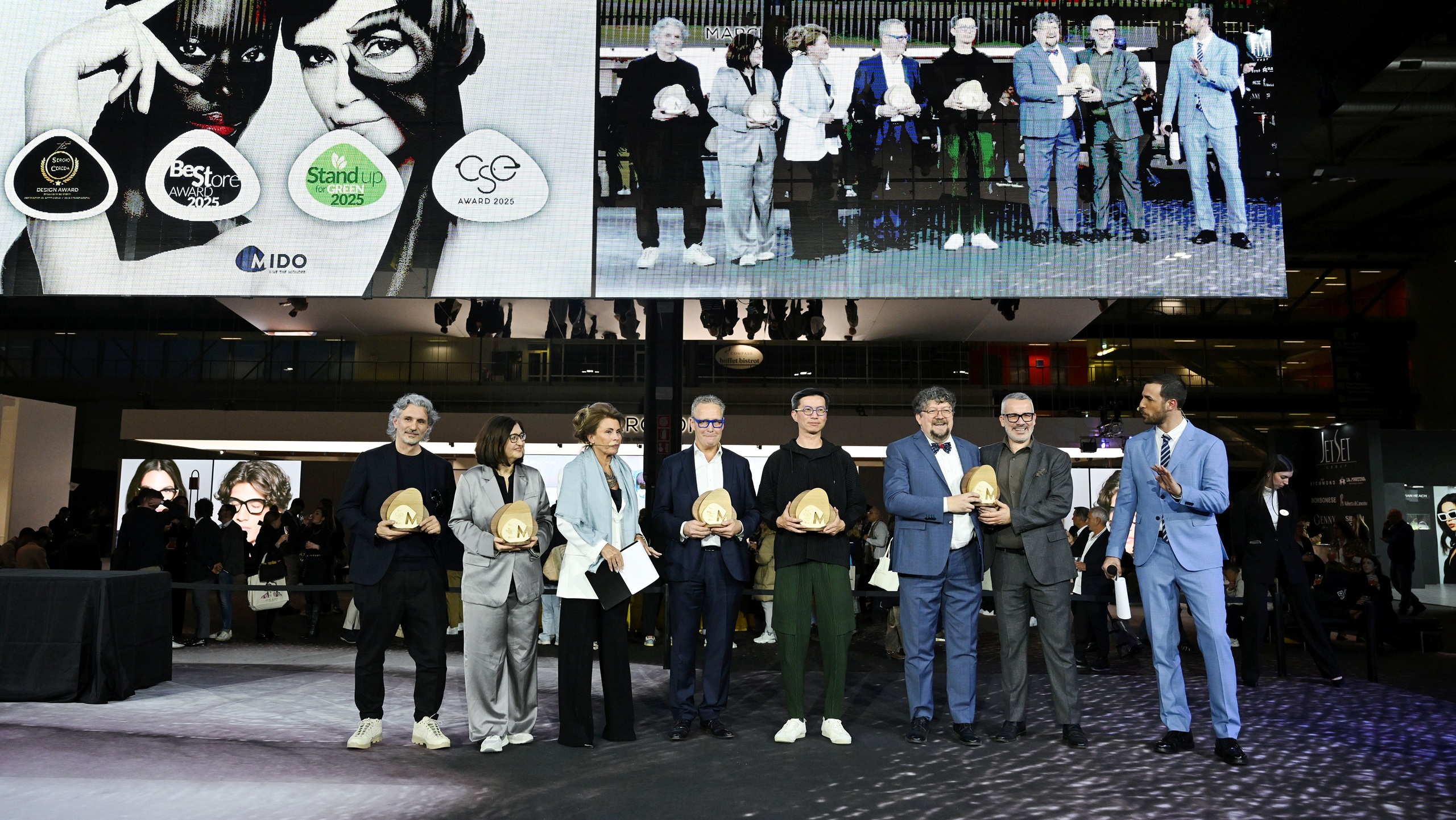 Cérémonie de remise des prix MIDO montrant un groupe de lauréats debout sur scène tenant des trophées dorés, avec un grand écran derrière eux affichant le branding de l'événement incluant les logos Best Store Award, Stand Up For Green 2025, CSE Award 2025, et MIDO aux côtés d'images de modèles portant des lunettes.