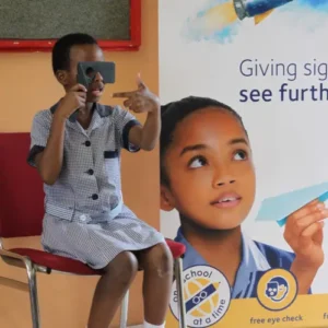 A young person in a striped shirt sits on a red chair while using a handheld vision screening device, pointing at something off-camera. Behind them is a large poster featuring a close-up of a child's face with text "Giving sight to see further" and logos including "free eye check" at the bottom