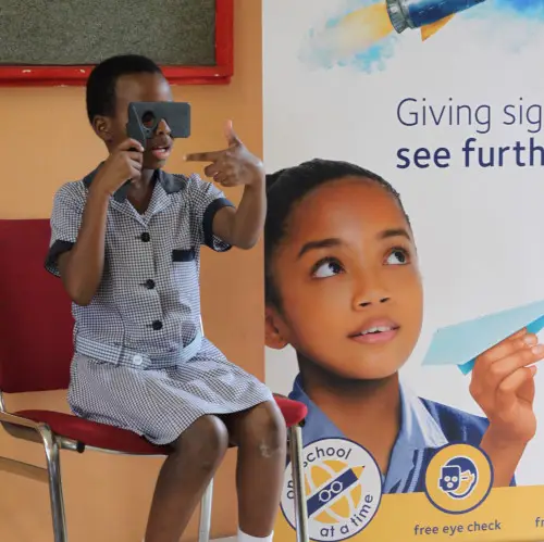 A young person in a striped shirt sits on a red chair while using a handheld vision screening device, pointing at something off-camera. Behind them is a large poster featuring a close-up of a child's face with text "Giving sight to see further" and logos including "free eye check" at the bottom