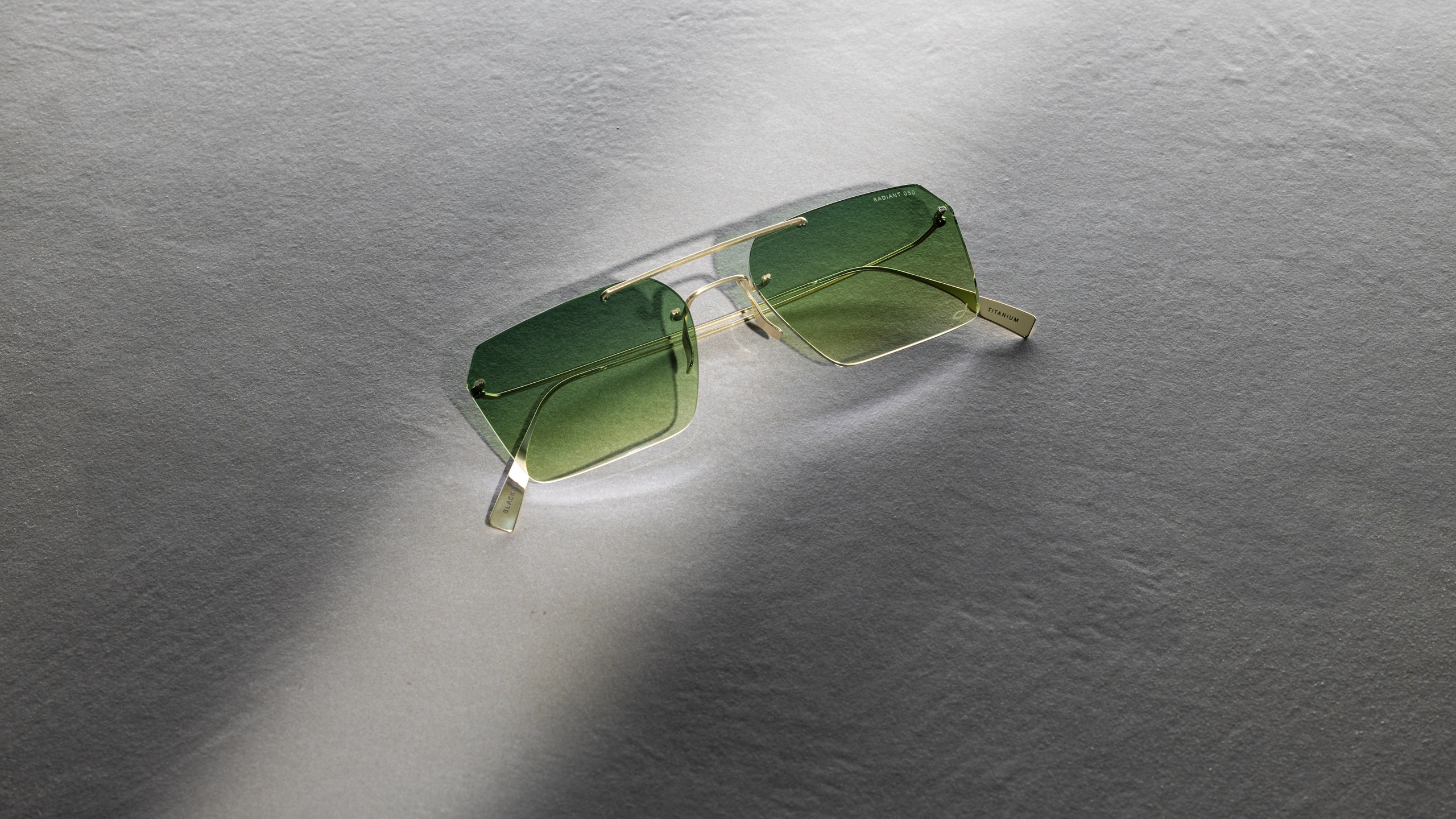 Lunettes de soleil Blackfin Aero Loop Infinity avec verres carrés géométriques et dégradé vert, présentant une construction en titane ultra-minimaliste avec design de pont géométrique, photographiées sur une surface grise texturée