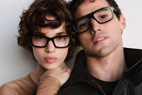 Two young people wearing black-framed eyeglasses posing closely together against a white background