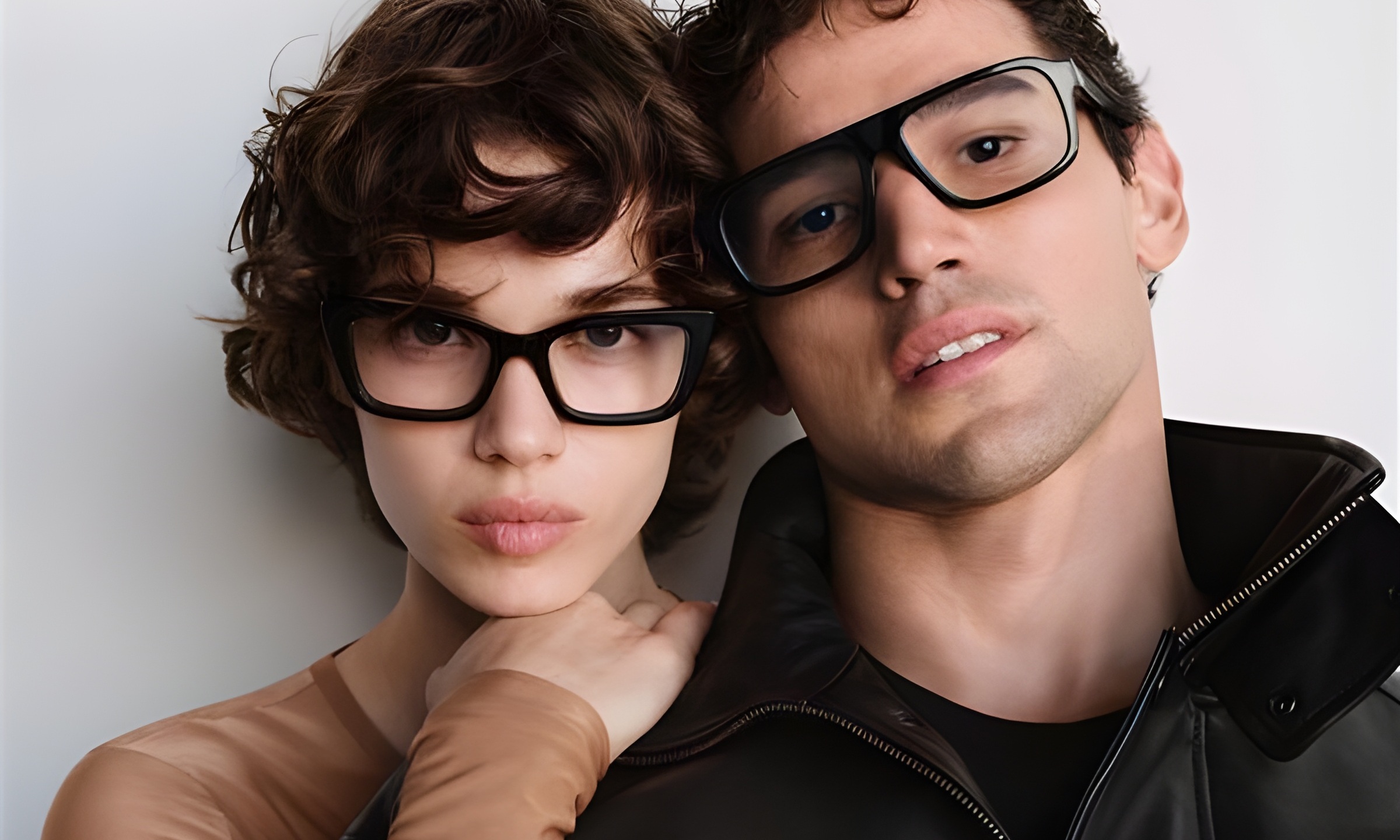 Two young people wearing black-framed eyeglasses posing closely together against a white background
