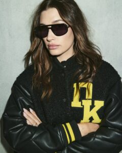 Hailey Bieber wearing black DKNY varsity jacket with yellow lettering and leather sleeves, paired with dark sunglasses, against industrial warehouse backdrop