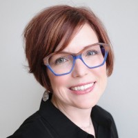 Portrait professionnel de Kelly Rosemann aux cheveux courts roux-brun, lunettes bleues et blazer noir souriant à la caméra