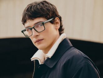 Mannequin masculin portant des lunettes optiques Lacoste à monture verte et des vêtements superposés marine et crème sur fond minimaliste.