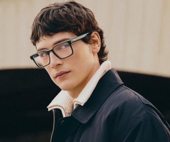 Mannequin masculin portant des lunettes optiques Lacoste à monture verte et des vêtements superposés marine et crème sur fond minimaliste.