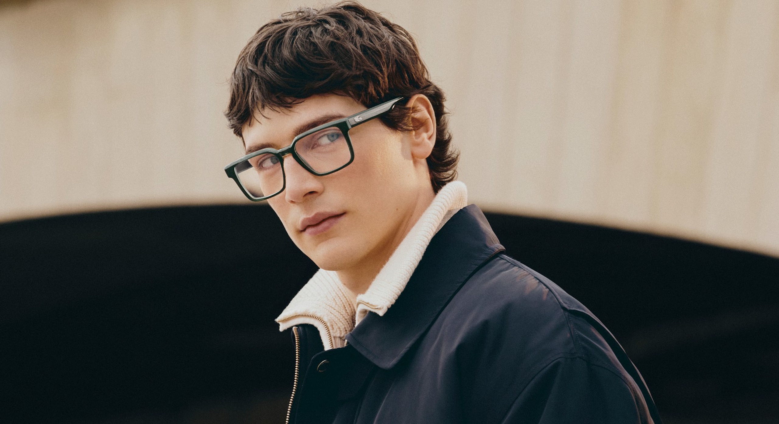 Mannequin masculin portant des lunettes optiques Lacoste à monture verte et des vêtements superposés marine et crème sur fond minimaliste.