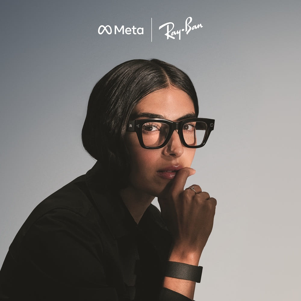 Portrait of a woman wearing black-framed Ray-Ban Meta smart glasses with her finger thoughtfully placed near her lips, with Meta and Ray-Ban logos visible above, emphasizing the blend of fashion and technology