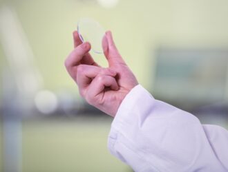Mains d'un professionnel en blouse blanche ou tenue médicale tenant une lentille transparente ou matériau optique pour l'examiner contre un arrière-plan de laboratoire ou clinique flou