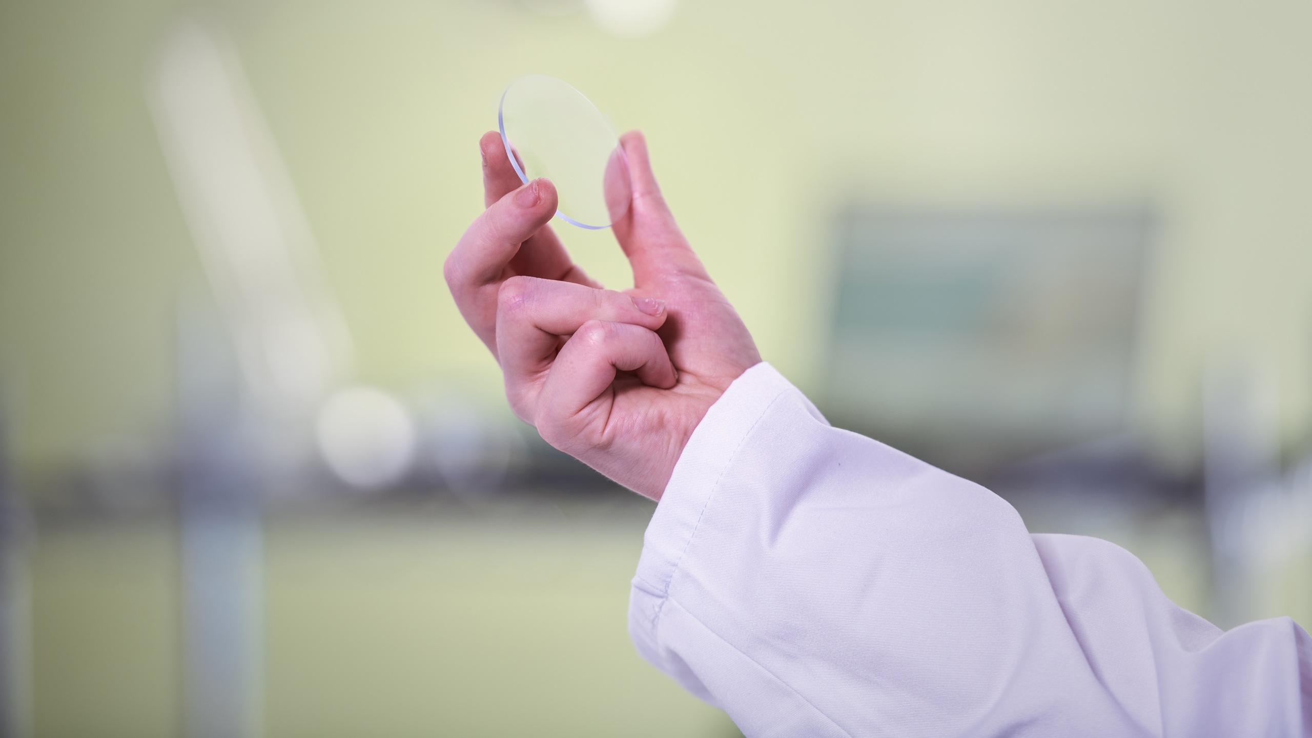 Mains d'un professionnel en blouse blanche ou tenue médicale tenant une lentille transparente ou matériau optique pour l'examiner contre un arrière-plan de laboratoire ou clinique flou