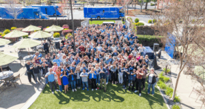 Aerial view of a large group photo with approximately 200-300 people gathered on a grassy area outside an office building. The crowd is densely packed and wearing a mix of business casual and casual clothing in various colors. Yellow and white market umbrellas are visible on the left side, along with blue trucks in the background parking area. The setting appears to be a corporate campus with landscaped areas, walkways, and bare winter trees surrounding the group.
