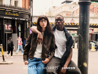 Image de campagne William Morris London présentant un jeune couple dans une rue animée de Londres. Une femme aux longs cheveux bruns portant des lunettes et une veste en cuir brun se tient avec un homme noir en t-shirt blanc et cardigan foncé. Derrière eux se trouve une scène de rue londonienne typique avec des bâtiments en brique rouge, des lampadaires noirs, et la devanture 'The Corner Shop Bar'. Le logo William Morris London apparaît en bas.