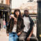 Image de campagne William Morris London présentant un jeune couple dans une rue animée de Londres. Une femme aux longs cheveux bruns portant des lunettes et une veste en cuir brun se tient avec un homme noir en t-shirt blanc et cardigan foncé. Derrière eux se trouve une scène de rue londonienne typique avec des bâtiments en brique rouge, des lampadaires noirs, et la devanture 'The Corner Shop Bar'. Le logo William Morris London apparaît en bas.