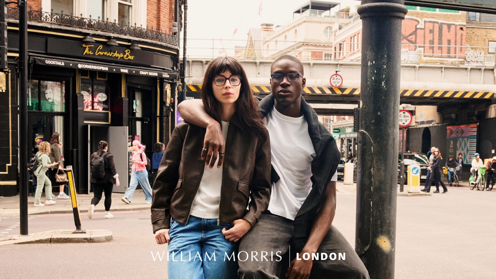 William Morris London eyewear campaign image featuring a young couple on a bustling London street. A woman with long dark hair wearing glasses and a brown leather jacket stands with a Black man in a white t-shirt and dark cardigan. Behind them is a typical London street scene with red brick buildings, black lamp posts, and 'The Corner Shop Bar' storefront. The William Morris London logo appears at the bottom.