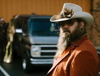Portrait en gros plan de Chris Stapleton portant un chapeau de cowboy blanc avec des détails de plumes, des lunettes de soleil foncées (lunettes Blackfin Eagle Head), et une veste de couleur rouille. Sa barbe longue distinctive est mise en évidence, avec des véhicules flous en arrière-plan, capturant une esthétique country classique.