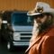 Portrait en gros plan de Chris Stapleton portant un chapeau de cowboy blanc avec des détails de plumes, des lunettes de soleil foncées (lunettes Blackfin Eagle Head), et une veste de couleur rouille. Sa barbe longue distinctive est mise en évidence, avec des véhicules flous en arrière-plan, capturant une esthétique country classique.