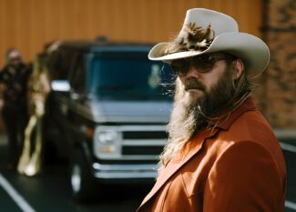 Close-up portrait of Chris Stapleton wearing a white cowboy hat with feather details, dark sunglasses (Blackfin Eagle Head eyewear), and a rust-colored jacket. His distinctive long beard is prominently featured, with vehicles blurred in the background, capturing a classic country aesthetic