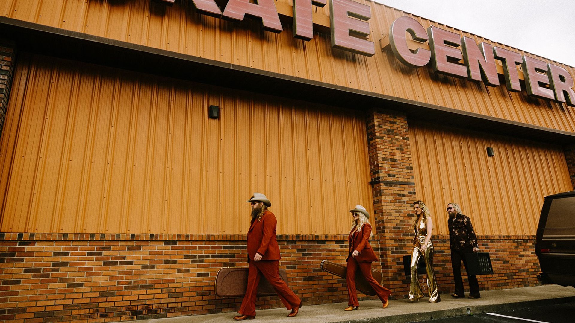 Quatre personnes en tenue western marchent le long du trottoir devant un bâtiment en brique avec une enseigne 'CENTER'. Deux silhouettes en costumes assortis de couleur rouille et chapeaux de cowboy mènent le groupe, suivies de deux autres en tenues contrastées, créant une scène stylisée rappelant la mode et la cinématographie des années 1970.