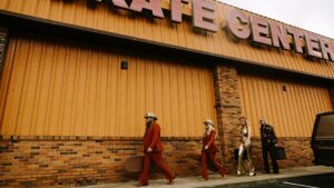 Four people in western attire walk along the sidewalk in front of a brick building with 'CENTER' signage. Two figures in matching rust-colored suits and cowboy hats lead the group, followed by two others in contrasting outfits, creating a stylized scene reminiscent of 1970s fashion and cinematography.