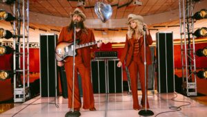 Chris Stapleton et Miranda Lambert performent ensemble sur une scène au style rétro, tous deux portant des tenues assorties de couleur rouille et des chapeaux de cowboy. Stapleton joue de la guitare tandis que Lambert se tient devant un microphone, avec un équipement d'éclairage vintage et une boule disco en forme de cœur visibles dans l'environnement de studio aux tons chauds.