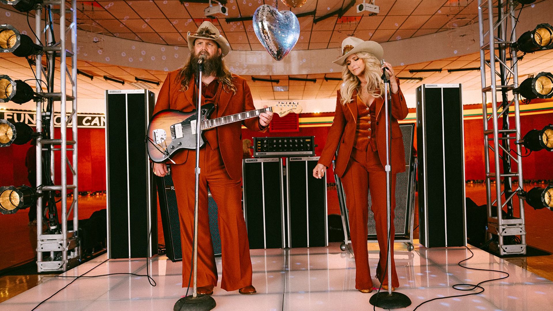 Chris Stapleton et Miranda Lambert performent ensemble sur une scène au style rétro, tous deux portant des tenues assorties de couleur rouille et des chapeaux de cowboy. Stapleton joue de la guitare tandis que Lambert se tient devant un microphone, avec un équipement d'éclairage vintage et une boule disco en forme de cœur visibles dans l'environnement de studio aux tons chauds.