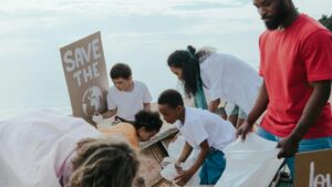 Un groupe de bénévoles diversifiés participe à une activité environnementale de nettoyage de plage. Plusieurs personnes portant des t-shirts blancs et colorés sont rassemblées pour ramasser les déchets et débris du sable, avec une personne tenant une pancarte "SAVE THE PLANET" illustrée d'une image de la Terre. La scène se déroule sur une plage avec les vagues de l'océan visibles en arrière-plan sous un ciel nuageux.