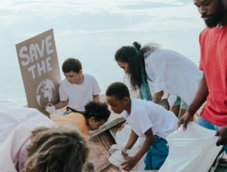 Un groupe de bénévoles diversifiés participe à une activité environnementale de nettoyage de plage. Plusieurs personnes portant des t-shirts blancs et colorés sont rassemblées pour ramasser les déchets et débris du sable, avec une personne tenant une pancarte "SAVE THE PLANET" illustrée d'une image de la Terre. La scène se déroule sur une plage avec les vagues de l'océan visibles en arrière-plan sous un ciel nuageux.