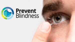 Close-up of a person's eye and finger as they insert or remove a contact lens, with the Prevent Blindness logo (featuring a circular design in blue, green, and yellow tones) displayed prominently in the upper left corner of the image.