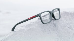 Lunettes de vue carrées Prada Linea Rossa en gris mat avec bande logo rouge, posées sur surface rocheuse glacée