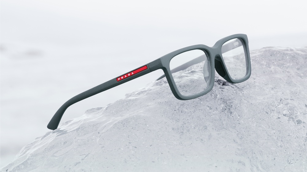 Lunettes de vue carrées Prada Linea Rossa en gris mat avec bande logo rouge, posées sur surface rocheuse glacée