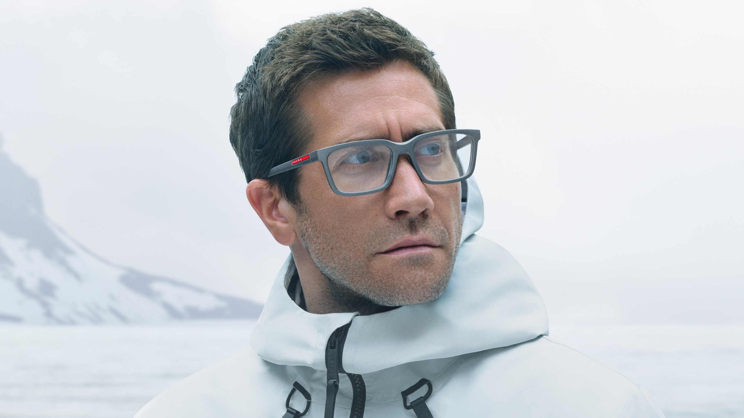 Jake Gyllenhaal wearing Prada Linea Rossa eyeglasses in white winter gear against misty mountain backdrop