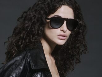 La mannequin Chiara Scelsi portant les lunettes de soleil Persol Total Black Arrow avec une veste en cuir noir, coiffée de cheveux bouclés volumineux sur fond gris atmosphérique