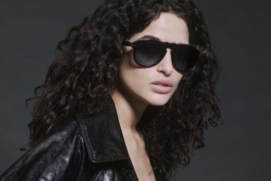 Model Chiara Scelsi wearing Persol Total Black Arrow sunglasses with black leather jacket, styled with voluminous curly hair against moody gray background