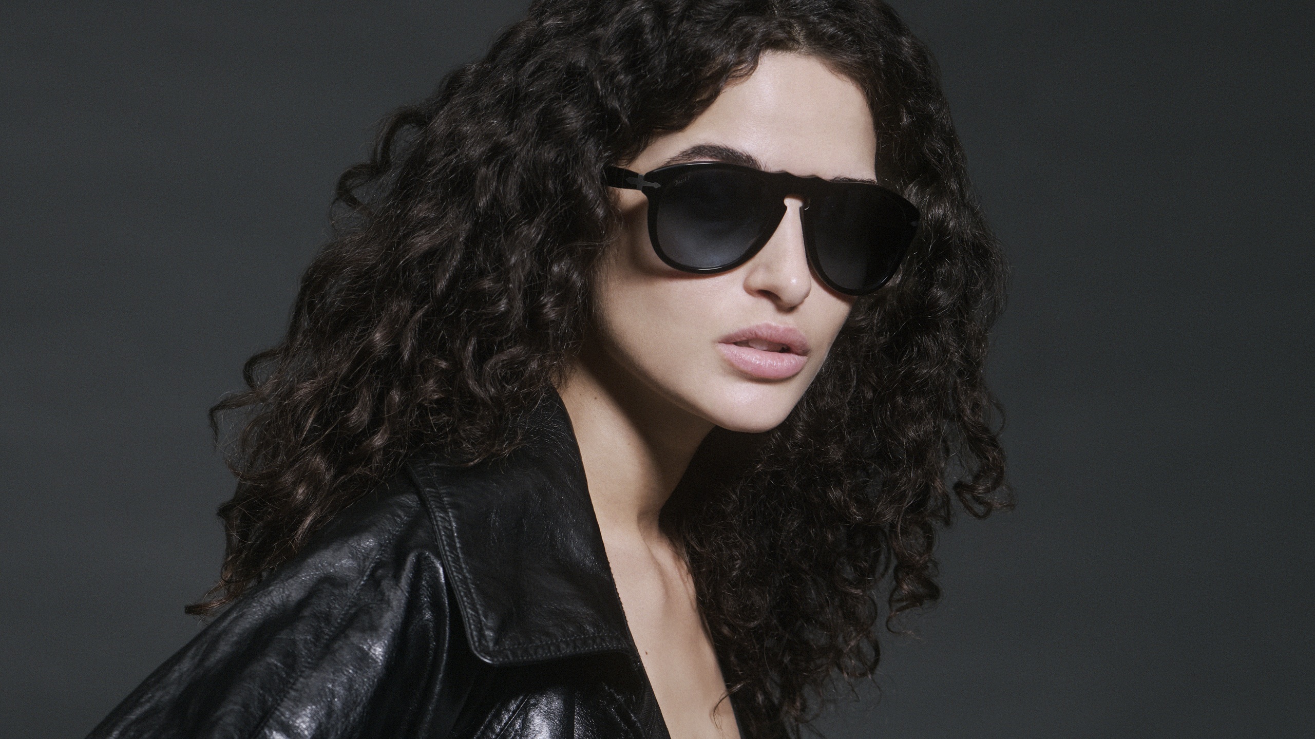 Model Chiara Scelsi wearing Persol Total Black Arrow sunglasses with black leather jacket, styled with voluminous curly hair against moody gray background