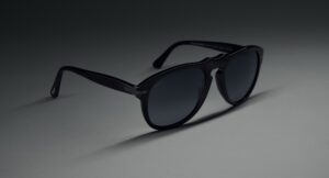 Persol 0PO0649NE Total Black Arrow sunglasses in classic pilot silhouette with distinctive arrow hinge detail, photographed at an angle on gray backdrop