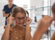 Young girl with braids trying on red eyeglasses at optometrist office