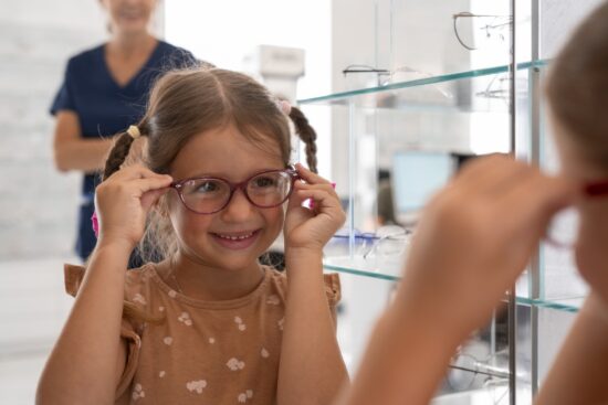 Young girl with braids trying on red eyeglasses at optometrist office