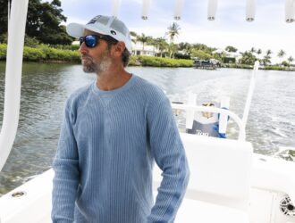 KC Scott portant des lunettes de soleil miroir bleues et une casquette blanche sur le pont d'un bateau avec des maisons au bord de l'eau visibles en arrière-plan
