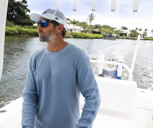 KC Scott portant des lunettes de soleil miroir bleues et une casquette blanche sur le pont d'un bateau avec des maisons au bord de l'eau visibles en arrière-plan