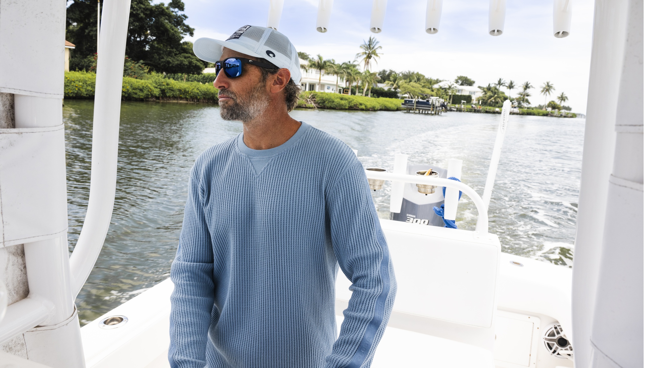 KC Scott portant des lunettes de soleil miroir bleues et une casquette blanche sur le pont d'un bateau avec des maisons au bord de l'eau visibles en arrière-plan