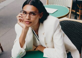 Woman wearing Lacoste L2316 optical glasses in a white blazer seated at a mint green café table in a modern Parisian setting