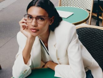 Woman wearing Lacoste L2316 optical glasses in a white blazer seated at a mint green café table in a modern Parisian setting